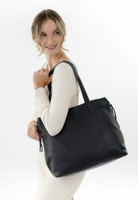 Sac fourre-tout en cuir noir avec une finition texturée, des poignées robustes et une fermeture à glissière, tenu par une personne portant un haut crème à côtes.