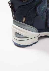 Botas de senderismo azul oscuro con un upper sintético liso, acentos de goma gris, correa de velcro y una suela texturizada que presenta una línea verde.