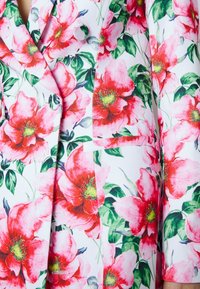 Blazer fleuri avec des fleurs rouges et roses vives sur un fond blanc. Fabriqué en tissu lisse, avec un col cranté et une poche avant.