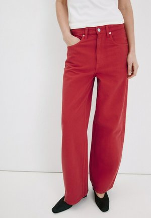 Mujer con pantalones rojos de tiro alto y pierna ancha con bolsillos frontales y zapatos negros puntiagudos, de pie frente a un fondo blanco liso.