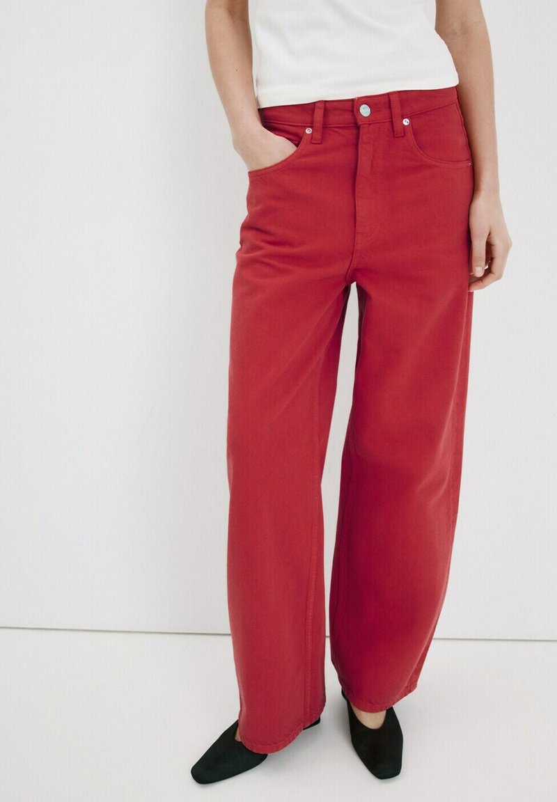 Femme portant un pantalon rouge taille haute à jambes larges avec des poches avant et des chaussures noires pointues, debout devant un fond blanc uni.