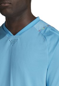 Lichtblauw sporttrui met een V-hals, textuurstof, geperforeerde details aan de voorkant en kleurrijke accenten op de schouder.