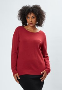 Pullover rosso con scollo a barca, maniche lunghe, orlo a coste e polsini decorati con piccole perline. Texture liscia.