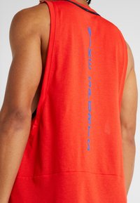 Rückansicht einer Person, die ein leuchtend rotes ärmelloses Oberteil trägt, auf dem "NIKE SPORTS" vertikal in Blau auf dem Rücken steht.