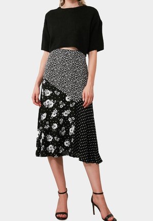 Frau trägt schwarzen kurz geschnittenen Pullover und schwarzen Midi-Rock mit Leoparden-, Blumen- und Polka-Dot-Mustern, kombiniert mit schwarzen hochhackigen Sandalen.