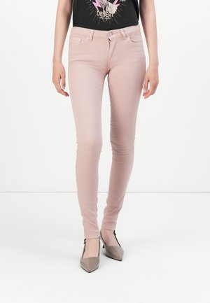 Kobieta nosząca jasnoróżowe obcisłe dżinsy w połączeniu z czarnym T-shirtem z nadrukiem i taupe szpiczastymi szpilkami, stojąca na białym tle.