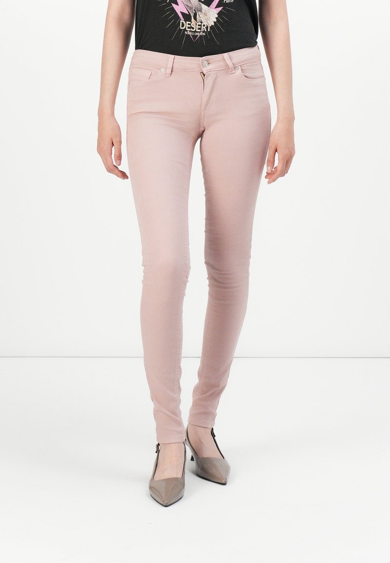 Femme portant un jean skinny rose clair associé à un t-shirt noir graphique et des talons pointus taupe, debout devant un fond blanc.