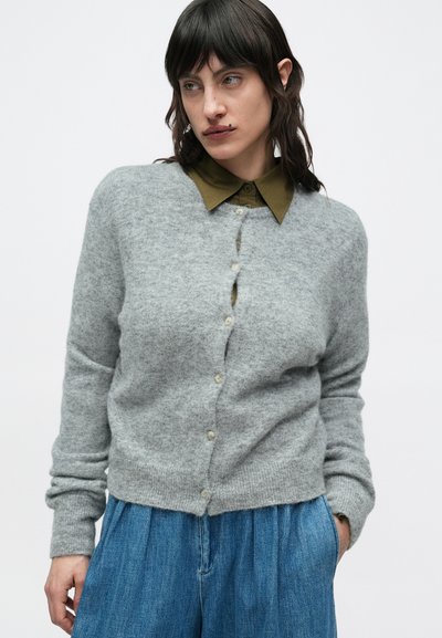 Γκρι μάλλινη ζακέτα με χαλαρή εφαρμογή, με στρογγυλή λαιμόκοψη, κλείσιμο με κουμπιά και ριμπ μανσέτες, φορεμένη πάνω από πουκάμισο με γιακά σε ελιά πράσινο χρώμα.