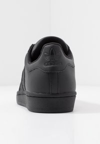 Černé kožené tenisky s texturovaným materiálem, mají černou gumovou podešev a vytlačené logo Adidas na patě. Jednoduchý, klasický design.