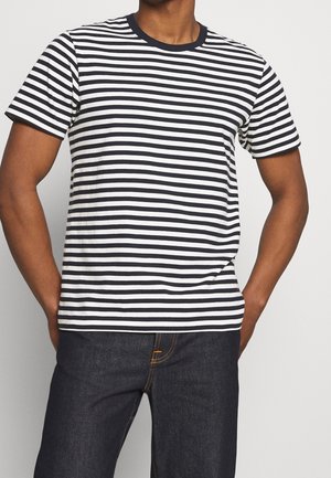 Hombre con camisa de manga corta a rayas horizontales negras y blancas y vaqueros azul oscuro, manos apoyadas en las caderas.