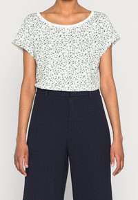 Vrouw in een witte bloemen kortemouwen top, gestoken in een hoge taille marineblauwe wijde broek, staande tegen een effen achtergrond.