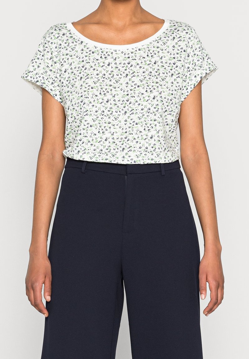Vrouw in een witte bloemen kortemouwen top, gestoken in een hoge taille marineblauwe wijde broek, staande tegen een effen achtergrond.