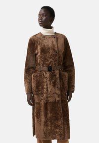 Hnědý střižový kabát s páskem v pase, který má velké přední kapsy a jemnou texturu, navržený s předním zapínáním na knoflíky.