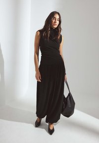 Črn brezrokavnik s cowl ovratnikom, v kombinaciji z ohlapnimi črnimi hlačami. Model drži veliko črno torbico in nosi črne čevlje s koničastim nosom.