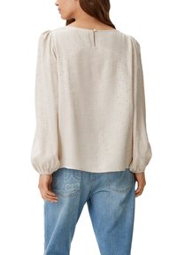 Helle beige Bluse mit Puffärmeln, subtiler Textur und Knopfverschluss hinten. Merkmale eines sich wiederholenden Textmusters in glänzender Ausführung.