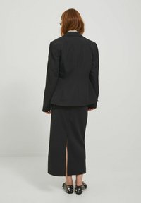 Blazer nero su misura con una forma aderente e colletto a punta, abbinato a una gonna coordinata con spacco alto. Le scarpe presentano dettagli scintillanti.