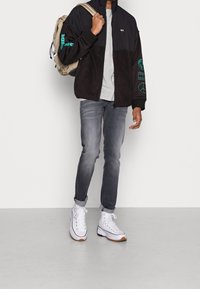 Černá a šedá bunda s barevnými bloky a tyrkysovými akcenty, šedé tričko, džíny středního odstínu a bílé vysoké tenisky. Batoh přehozený přes rameno.