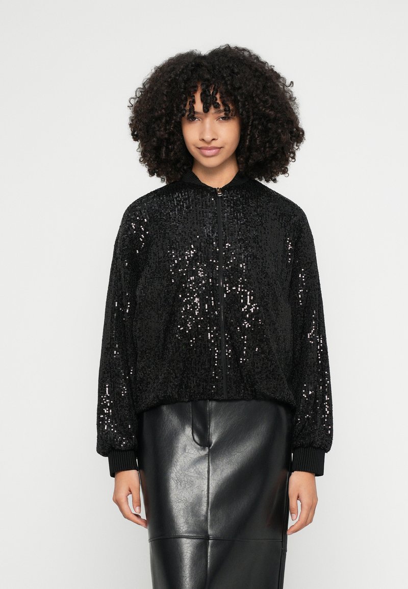 Juoda bombonešio striukė, padengta juodomis blizgučiais, su priekyje esančia užtrauktuku ir austo audinio rankogaliais. Dėvima su juoda odine sijonu.