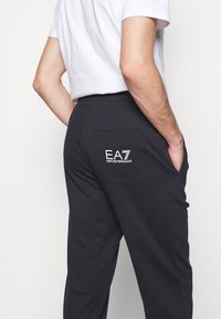 Černé bavlněné tepláky s kapsou vzadu, s bílým logem EA7 Emporio Armani. Elastický pas, klasický střih, hladká textura.