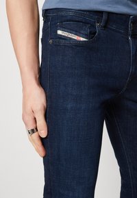 Vaqueros de mezclilla azul oscura con una textura suave, diseño de cinco bolsillos y una etiqueta de marca visible en el bolsillo trasero. Acentos de hardware plateado.