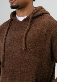 Hoodie polaire marron texturé avec cordons de serrage et capuche, présentant une surface douce et duveteuse. Vue en gros plan montrant les détails du tissu.