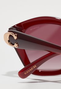 Γυαλιά ηλί�ου με κόκκινη απόχρωση και χοντρό σκελετό, εφοδιασμένα με χρυσή έμβλημα στον βραχίονα και με την ένδειξη "COACH" χαραγμένη στην αγκαλιά. Γυαλιστερή επιφάνεια.