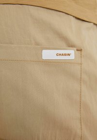 Béžové látkové šortky s hladkou texturou; mají zadní kapsu s bílou štítkem s nápisem "CHASIN'" hnědým písmem.