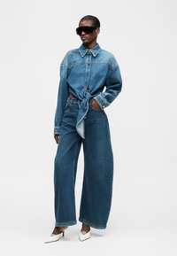 Naine, kes kannab suurt seotud teksasärki, laiade sääreosadega teksapükse, suuri musti päikeseprille ja valgeid teravate kontsadega kingi, seisab ühevärvilise tausta ees.