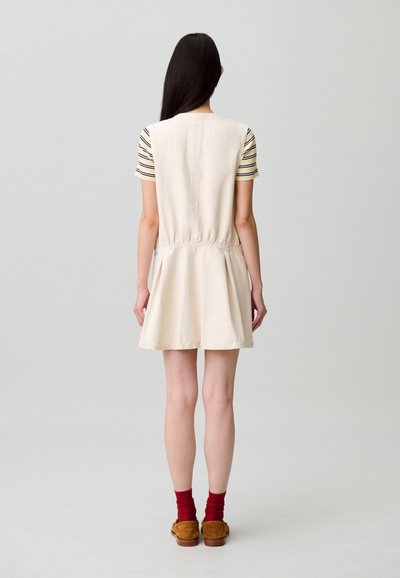 Vestido beige claro con cintura ceñida, falda acampanada y mangas cortas. Capa inferior de manga corta a rayas y zapatos marrones con calcetines rojos.