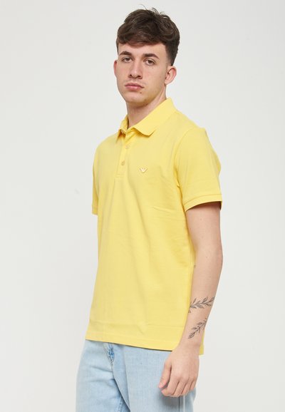 Polo gialla in cotone con taglio dritto, maniche corte e un piccolo logo sul lato sinistro del petto. Indossata con jeans azzurri chiari.