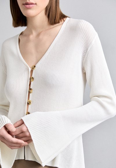 Λευκό ριγωτό ζακέτα με V-λαιμόκοψη, διαθέτει πέντε χρυσά κουμπιά μπροστά και φαρδιά μανίκια, φτιαγμένη από μια υφή ύφασμα.