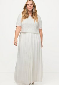 Abito lungo bianco con top a maniche corte decorato con perline. La silhouette fluida e il tessuto testurizzato creano un aspetto elegante. Accenti argentati visibili.