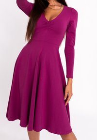 Robe ajustée à manches longues en magenta vif, avec un col en V et un détail froncé au niveau de la poitrine, s'évasant en une jupe longueur genoux.