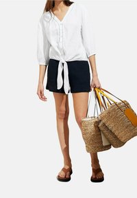 Blusa bianca in lino con un centro annodato, abbinata a pantaloni corti texturizzati blu scuro. La modella porta tre borse intrecciate di diverse dimensioni.