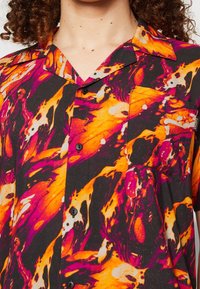 T-paita lyhyillä hihoilla, mustalla taustalla, jossa on abstrakti kuvio oranssin, vaaleanpunaisen ja valkoisen kieppuvan kuvion muodossa. Sisältää rintataskun ja napit.