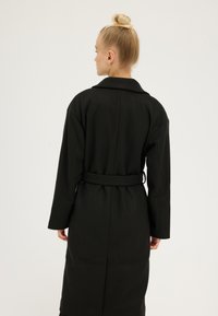 Woman with blonde hair in a bun wearing a long black belted coat, standing facing away against a plain white background.