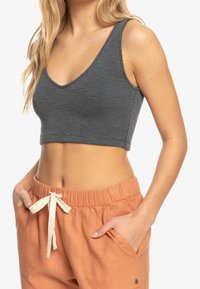 Kvinde iført en ærmeløs grå crop top og højtaljede, løse orange bukser med en hvid snor, hænderne i lommerne.