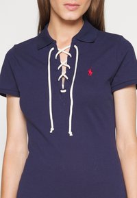 Námořnická polo tričko s designem v-neck se šněrováním, s krémovými šňůrkami a červeným vyšitým logem na hrudi. Hladká textura.