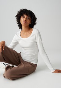 Woman with curly hair sitting on floor, wearing white long-sleeve top, brown pants, and white sneakers against plain gray background.