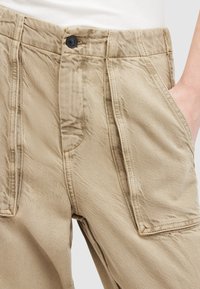 Pantalon décontracté beige avec poches à rabat à l'avant et fermeture à bouton noir, porté avec un haut blanc rentré et une main dans la poche.