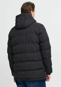 Svart pufferjakke med hette, med quiltet design, strukturert stoff og ribbet søm, vist fra baksiden.