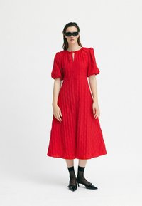 Rochie midi roșie cu un model texturat, mâneci bufante și un detaliu legat la guler. Purtați cu pantofi și șosete negre.