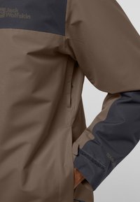 Bruine waterdichte jas met marineblauwe accenten, met een soepele textuur, logo-opdruk op de borst en klittenbandsluitingen bij de mouwen.