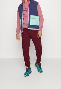 Fleece vest v námořnické modři s mentolovými akcenty a růžovým zipem, vrstvený přes bordó mikinu a odpovídající tepláky. Tyrkysové a černé sportovní boty.