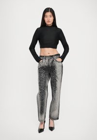 Woman wearing black cropped long-sleeve top, high-waisted faded black and gray patterned jeans, and black pointed heels, standing with hands in pockets.
