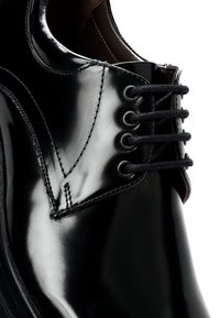 Zapato de vestir de cuero negro con un acabado brillante, que presenta siete ojetes, cordones negros y detalles de costura sutiles a lo largo de la punta redondeada.