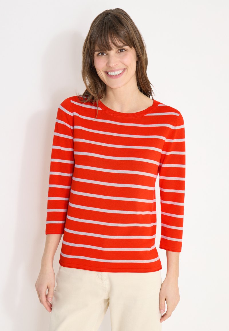 Femme souriante portant un pull rouge à rayures horizontales blanches et un pantalon beige clair, debout devant un fond blanc uni.