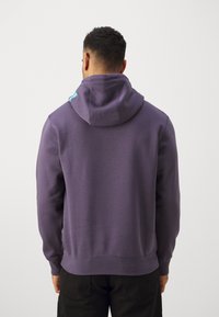 Felpa viola con una vestibilità rilassata e cappuccio con coulisse. Presenta un accento blu sulla spalla, realizzata in morbido cotone.