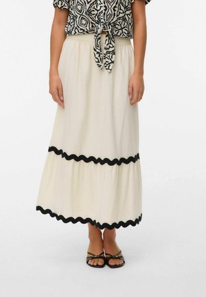 Kvinde iført en cremet ankel-lang nederdel med sort bølget kant, kombineret med sorte remsandaler og en mønstret knyttet bluse.