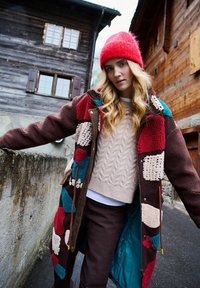 Žena v farebnom patchworkovom kabáte, s červenou pletenou čiapkou a béžovým svetríkom stojí na úzkej ulici s drevenými domami v pozadí.